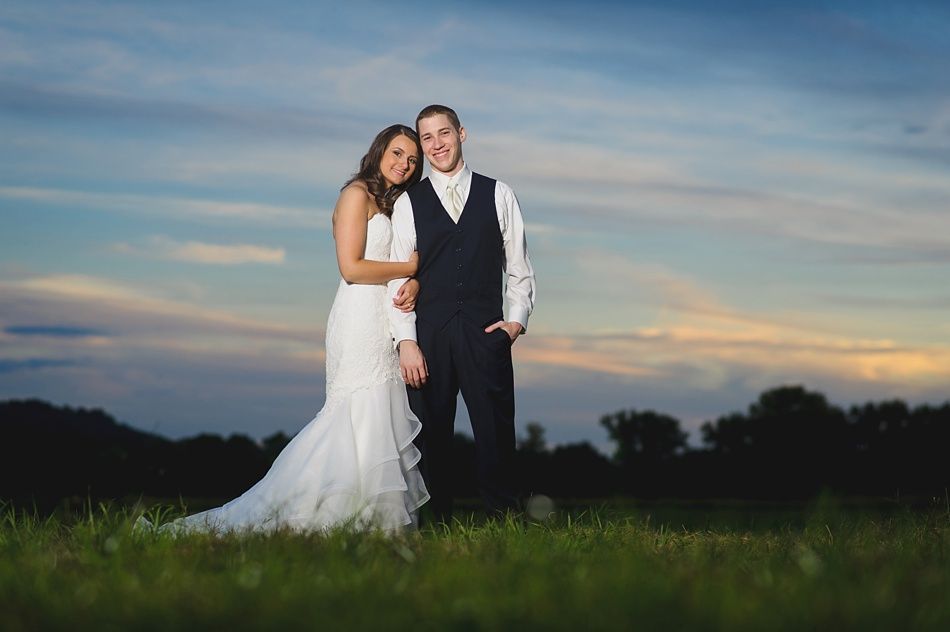 Couple in wedding attire pose in a field at dusk; woman leans on man, sunset colors in the sky.