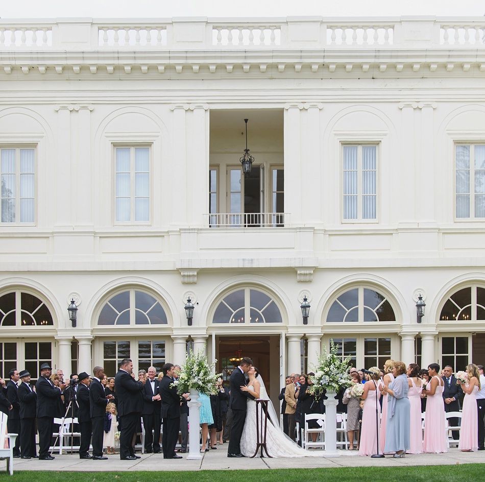 Wedding ceremony outside a white mansion; couple kissing, guests in formal attire.