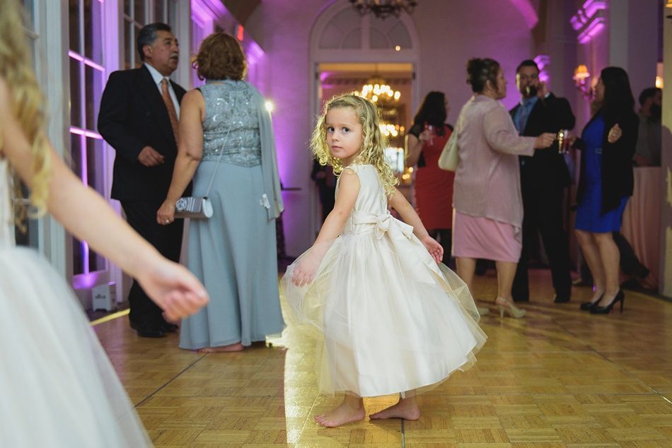Young girl in a cream dress dances on a wooden floor at an event, other guests in background.