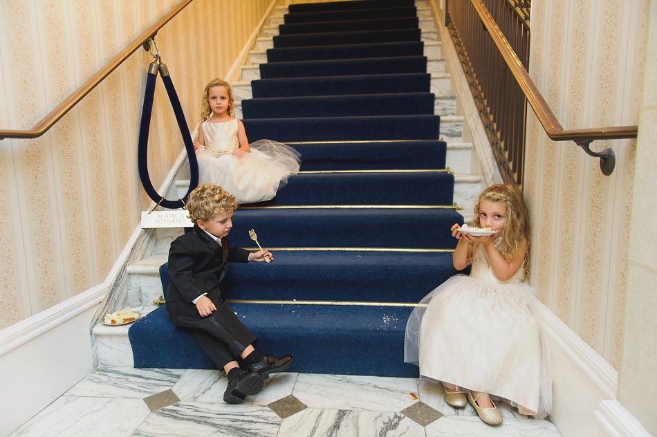 Three children, two girls in white dresses, one boy in a suit, on a staircase with blue carpet. They are eating cake.