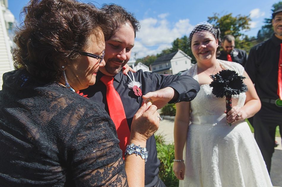 Bride and groom exchange rings, assisted by a woman in a black lace top. Outdoors, sunny. Groom in red tie, bride in white dress.