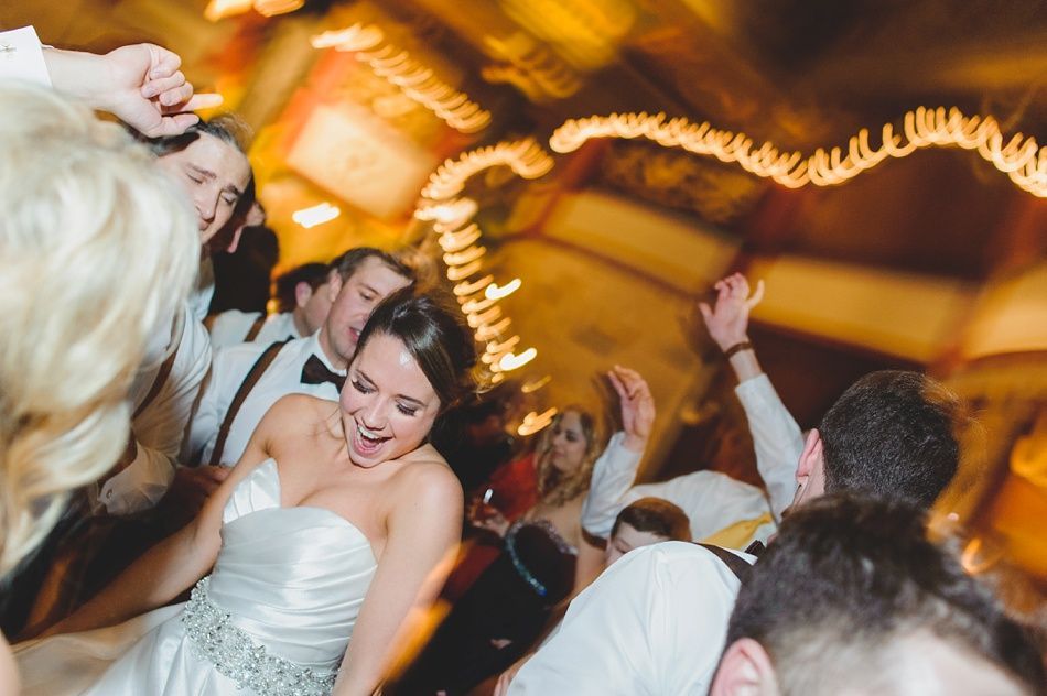 Bride dancing at reception, smiling, surrounded by party guests; festive lights in background.