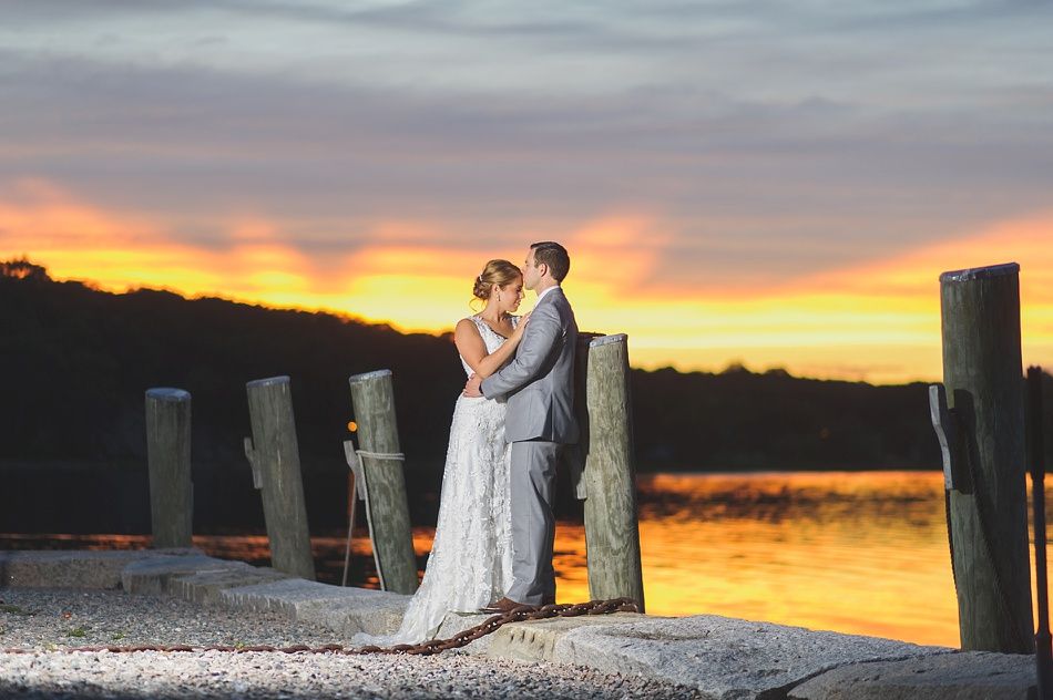 Couple embracing on a dock at sunset; orange and yellow sky reflecting on the water.
