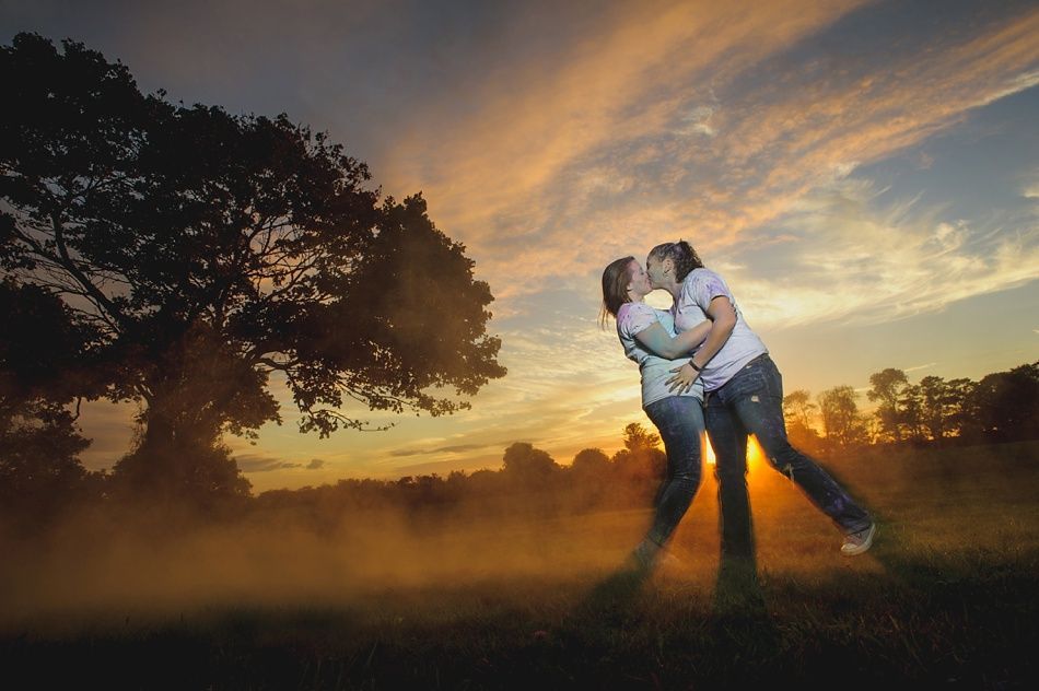 Two people kissing at sunset; silhouetted tree, colorful sky, hazy field.
