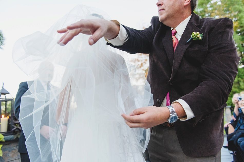 Father lifting bride's veil during outdoor wedding ceremony. Groom, attendees, and candles in the background.