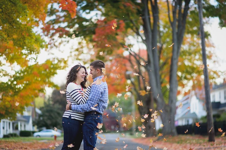 Couple embracing on a street with falling autumn leaves, smiling at each other. Colorful trees in background.