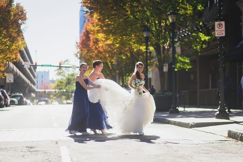 Bride in white dress, two bridesmaids in blue dresses, crossing a city street. Autumn leaves and sunlight.