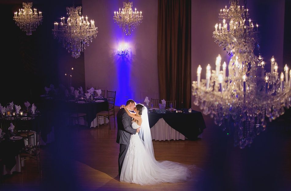 Bride and groom embrace on a dance floor, surrounded by chandeliers and tables set for a wedding.