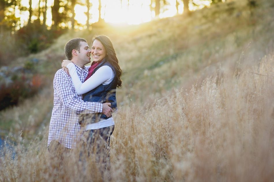 Couple embraces in a field, smiling at each other. Sunlight illuminates the background.