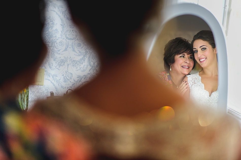 Bride and mother in a mirror; mother smiles, bride looks radiant in dress.