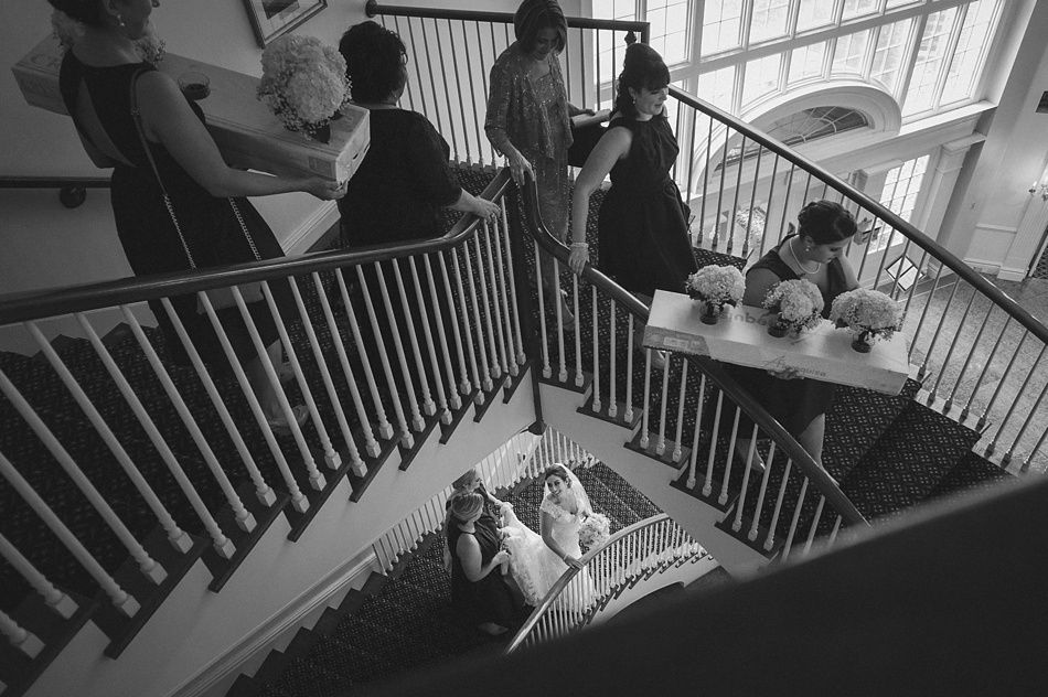 Wedding party on a grand staircase. Bridesmaids carry bouquets. Bride and another look up. Black and white photo.