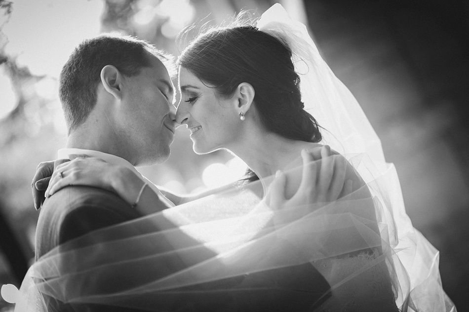 Bride and groom embrace, nearly kissing, outdoors. Bride's veil is billowing in sunlight. Black and white.