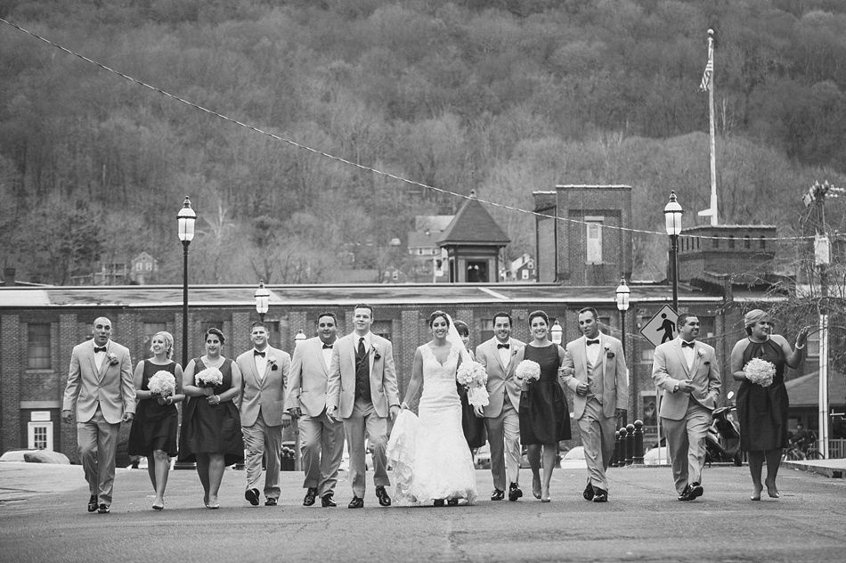 Wedding party walking down a street in front of a brick building and forested hill. Black and white.