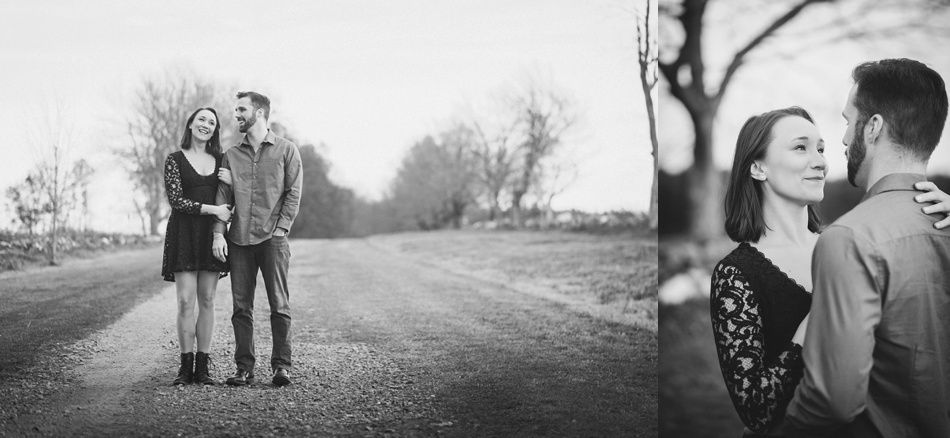 Couple on a gravel road, woman in a black dress, man in a button-down shirt. Embracing, smiling. Black and white.