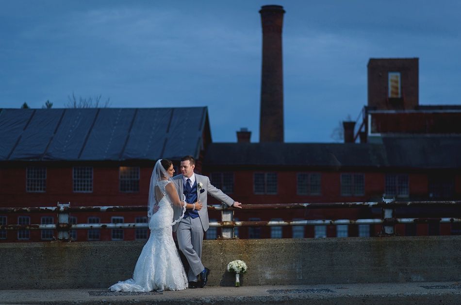 Bride and groom pose by a red brick building at dusk. Bride in white dress, groom in grey suit, facing each other.
