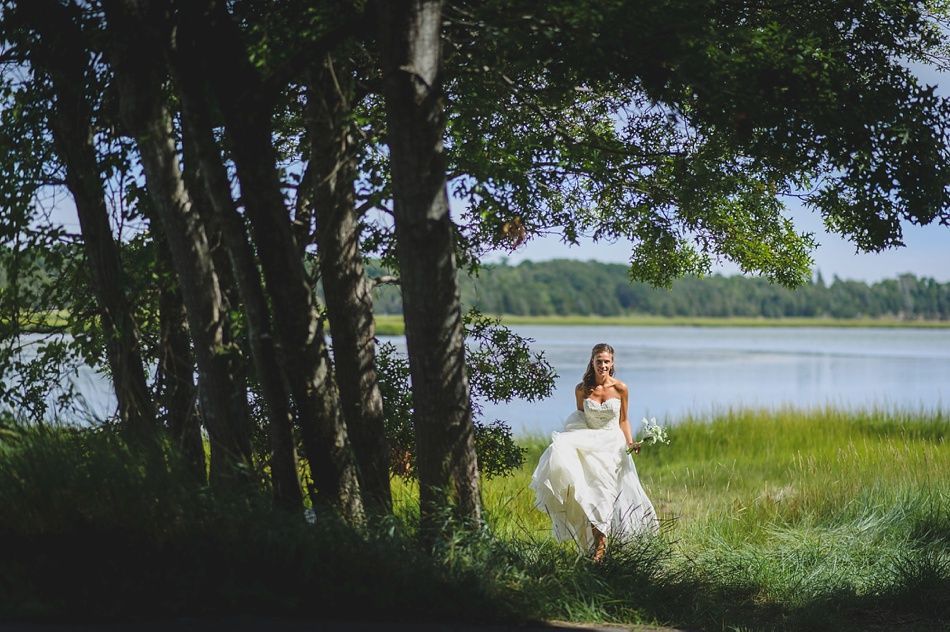 Bride in a white gown stands near a lake, holding a bouquet, under the shade of trees.