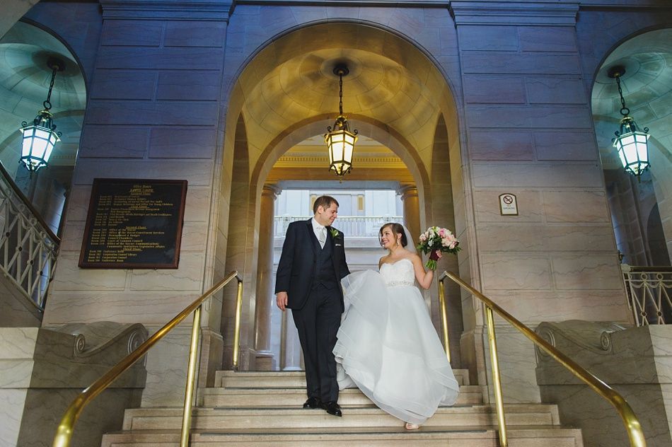 Couple in wedding attire descends stairs under an archway. Gold railings, stone walls, and hanging lanterns.