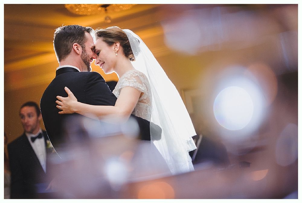 Couple dancing at wedding, man in tuxedo, woman in white dress and veil, blurred background.