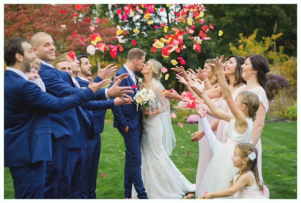 Wedding party celebrating couple's kiss with tossed rose petals outdoors. People in blue and pink outfits smile.