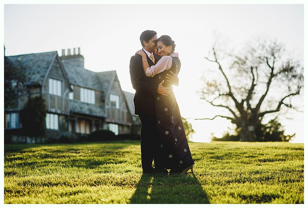 Couple embracing on a grassy lawn with a large house in the background; sunlight streams through a bare tree.