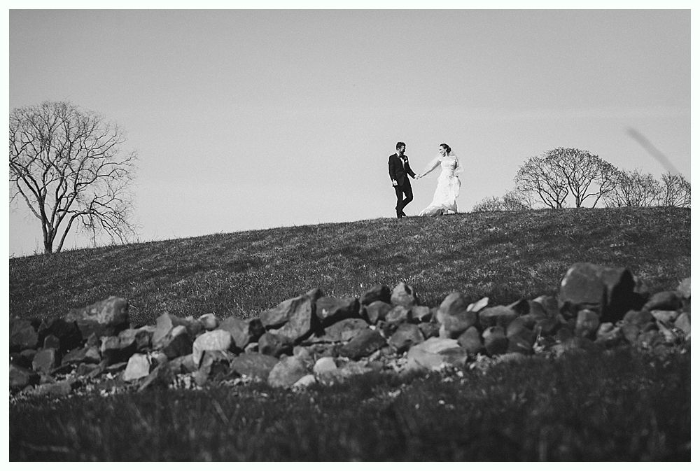 Couple holding hands, walking on a hilltop. Wedding attire, black and white photograph, trees in the background.