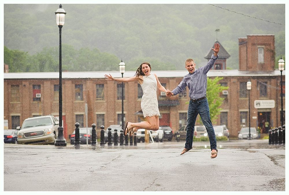 Couple jumping joyfully in front of brick building on a rainy day, holding hands, arms outstretched.