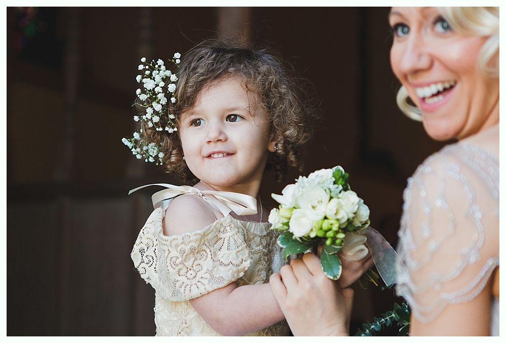 A young girl in a lace dress smiles, holding flowers with a smiling woman nearby.