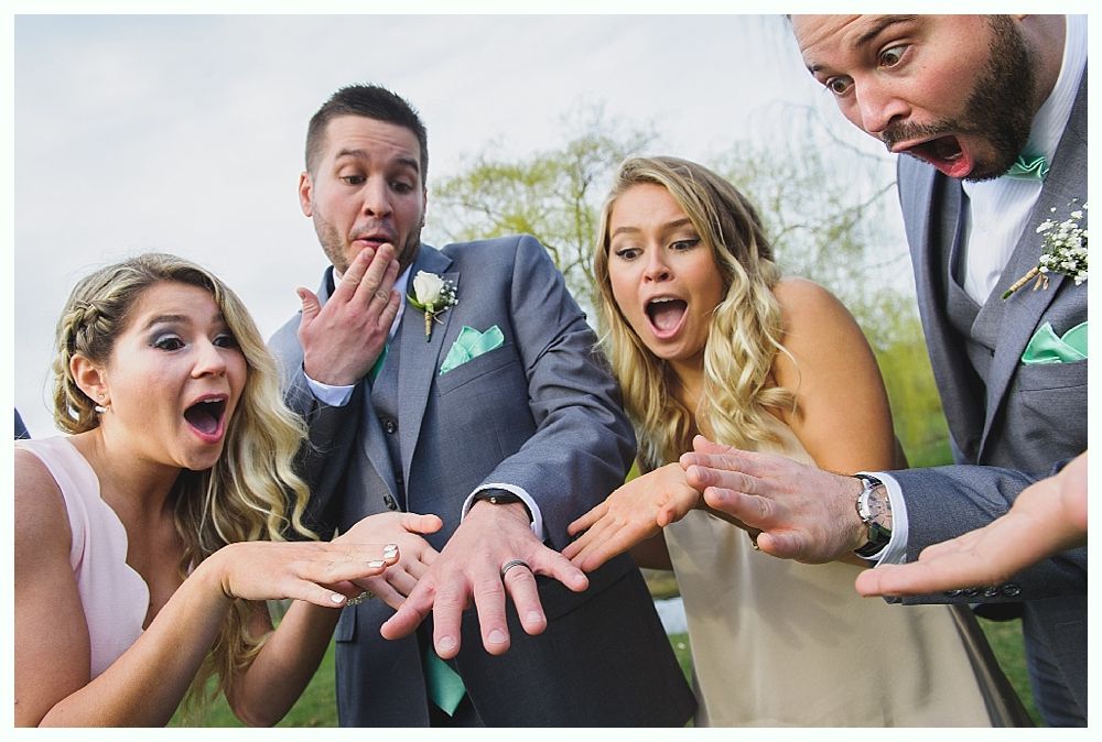 Four people with surprised expressions, looking at a wedding ring on a man's hand. Outdoors, sunny day.