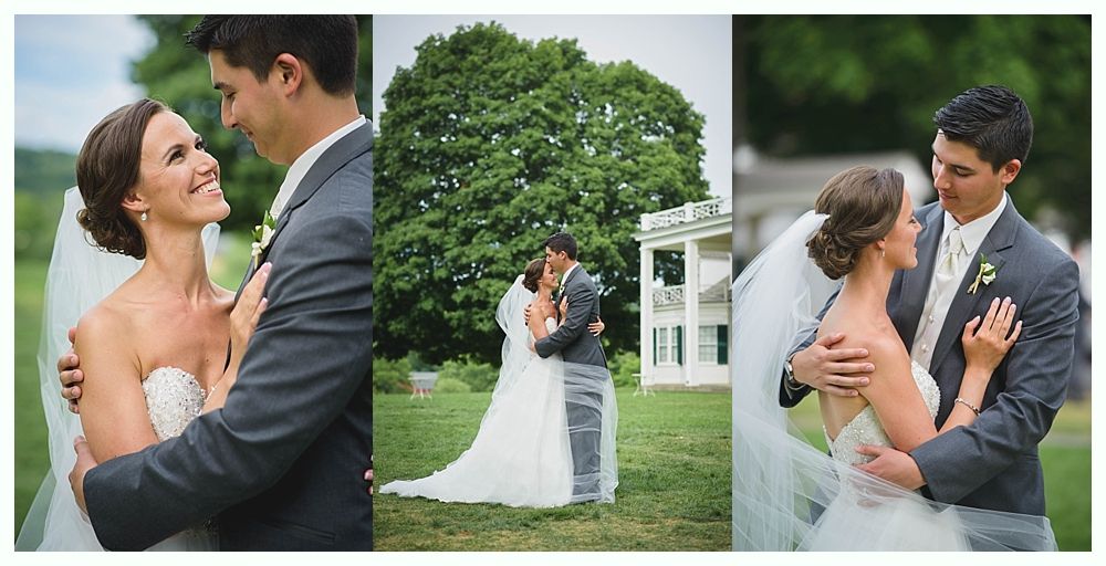 Wedding couple embraces in a grassy field. Bride in strapless dress, groom in grey suit, with a large tree and white building in the background.