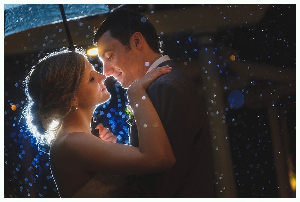 Couple embracing on a beach, smiling; man in pink shirt, woman in white, wooden railing, sunny day.