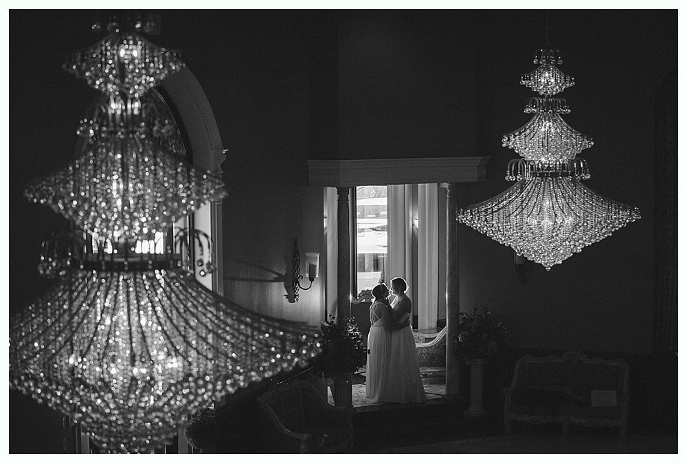 Two brides embracing in a doorway, framed by ornate crystal chandeliers. Black and white.
