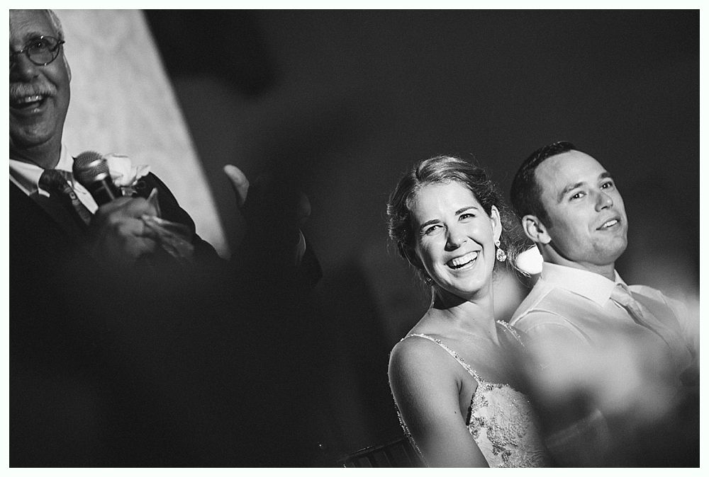 Bride and groom laughing during a wedding toast. Man with microphone in background.