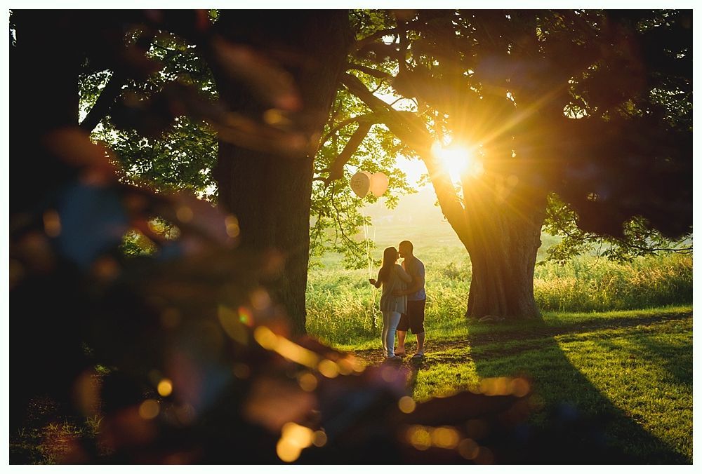 Couple standing in a sunlit park with balloons, sunlight streaming through trees.