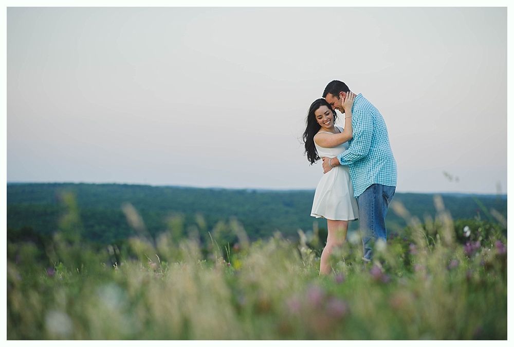 Couple embracing in a field with tall grass, man in blue shirt, woman in white dress, smiling.