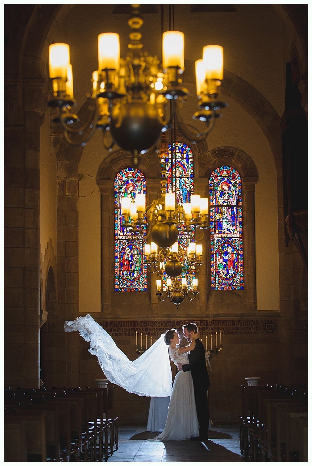 Couple kissing under chandelier in church, bride's veil floating, stained glass windows.