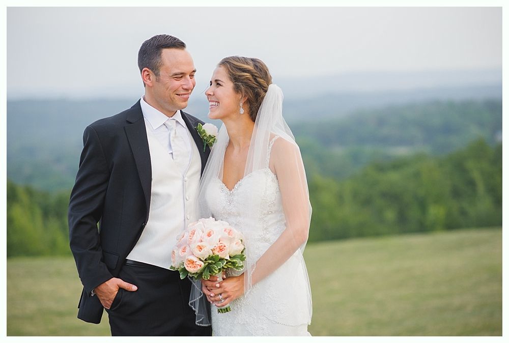 Bride and groom looking at each other outdoors, holding bouquet. She wears a white dress, veil; he wears a suit. Green hills in background.