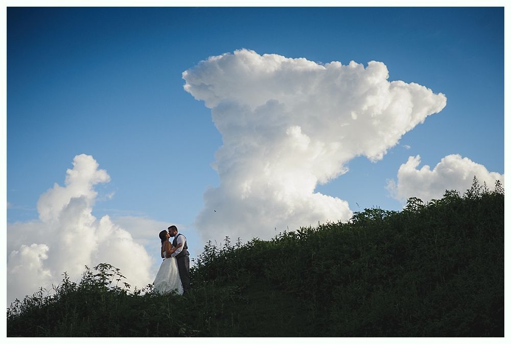 Couple kissing on a grassy hill beneath a large, white cloud in a blue sky.