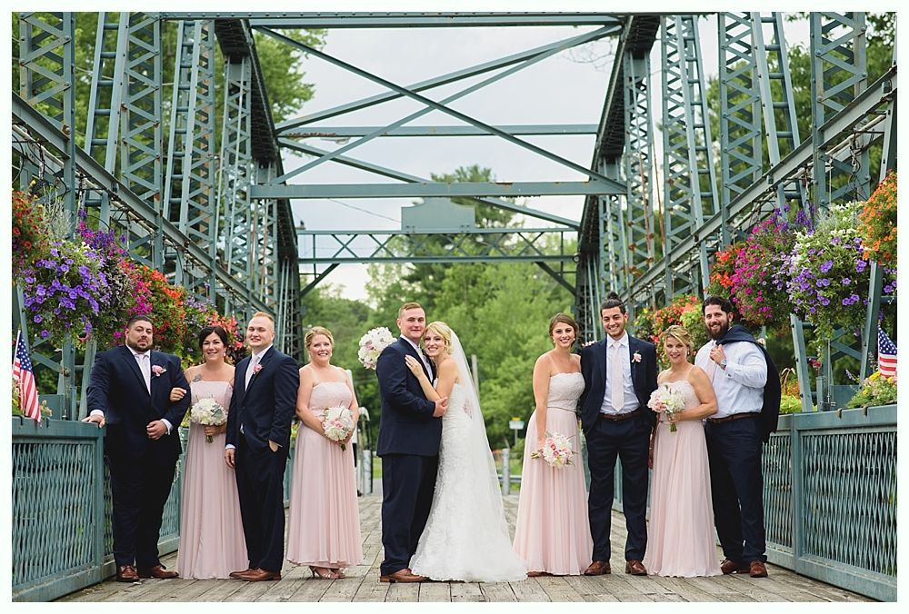 Wedding party on a green bridge, bride and groom in the center, bridesmaids in pink, groomsmen in navy, flowers.