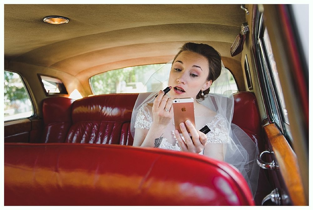 Bride in vintage car applying lipstick, looking in phone screen, wearing veil.