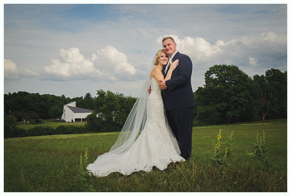 Newlyweds embrace in a field with a white building and trees in the background, cloudy sky.