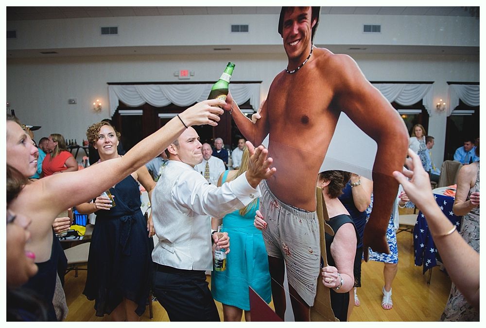 Guests celebrate a wedding. A shirtless cutout figure holds a beer; guests reach for it.