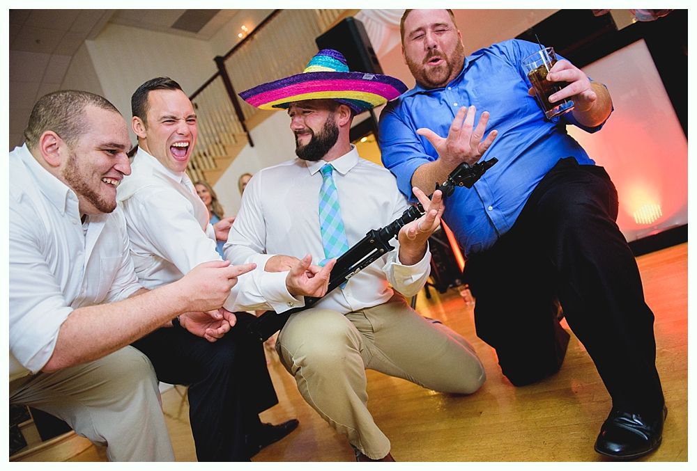 Men at a party, one in a sombrero playing a pretend instrument, others laughing.
