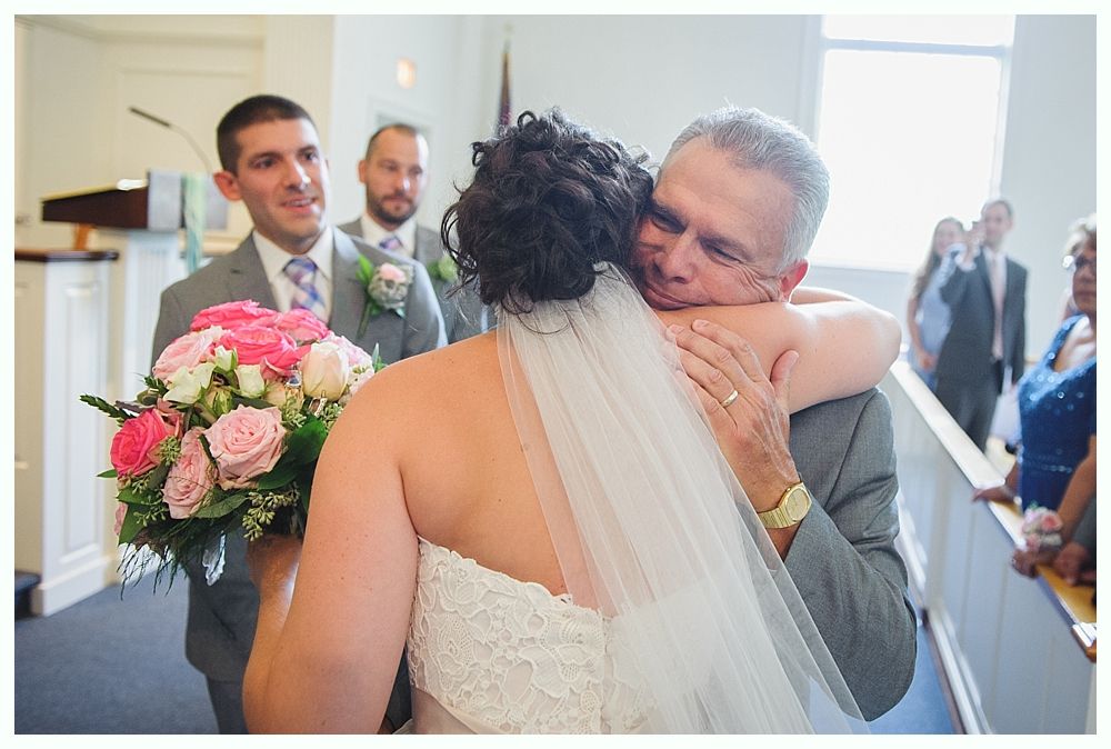 Bride in white dress embraces a man in gray suit at a wedding ceremony.