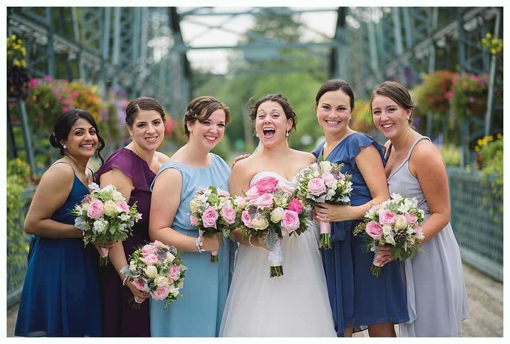 Bride and bridesmaids smiling on a bridge, holding bouquets. Dresses in various shades of blue and purple.