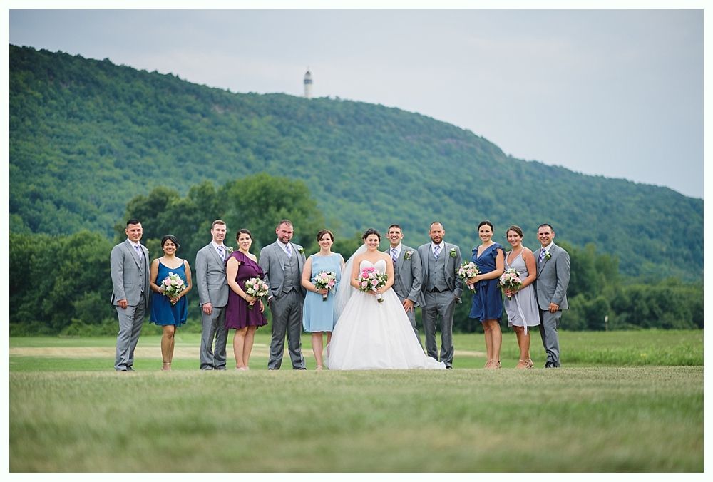 Wedding party posing on a grassy field with a mountain and lighthouse in the background.