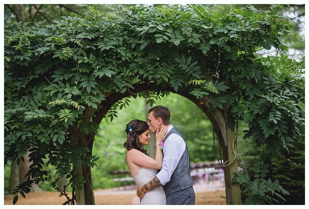 Couple embraces under a leafy archway outdoors; the man kisses the woman's forehead, surrounded by greenery.