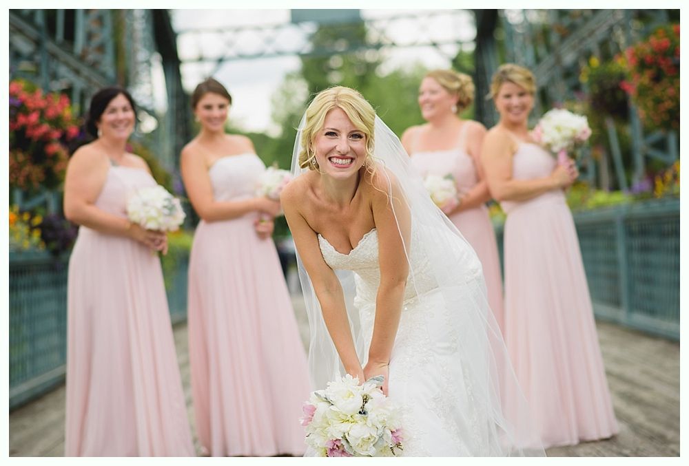 Bride in white gown and veil smiles, surrounded by bridesmaids in pink dresses on a bridge, holding bouquets.
