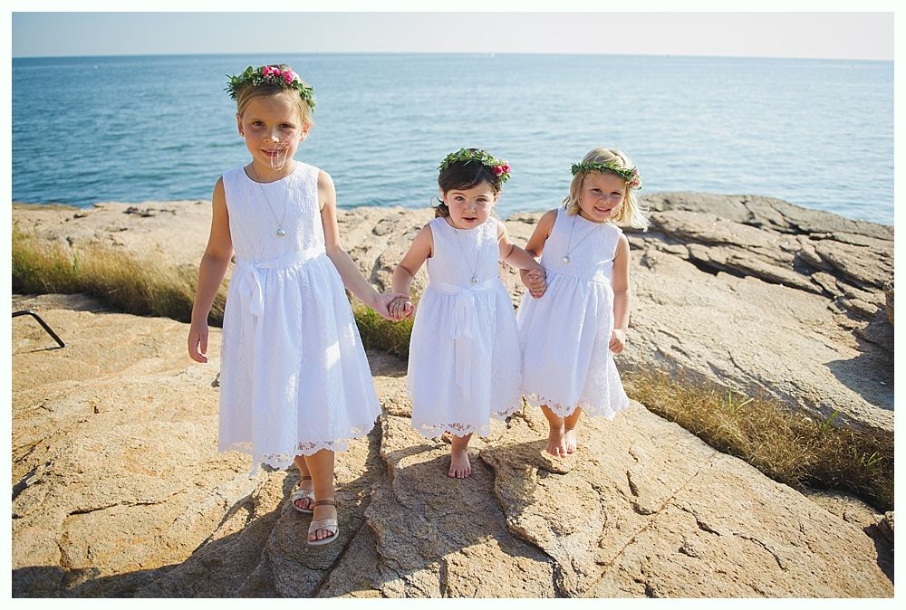 Three young girls in white dresses holding hands, wearing flower crowns, on a rocky beach by the ocean.