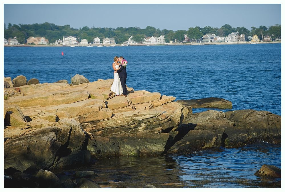 Couple embraces on rocky shore, ocean and town in background. Sunny day.