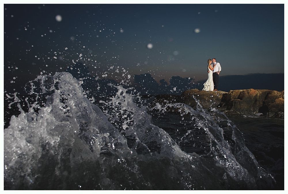 Newlyweds embrace on rocks as waves crash. Ocean at dusk, dark sky, white dress.